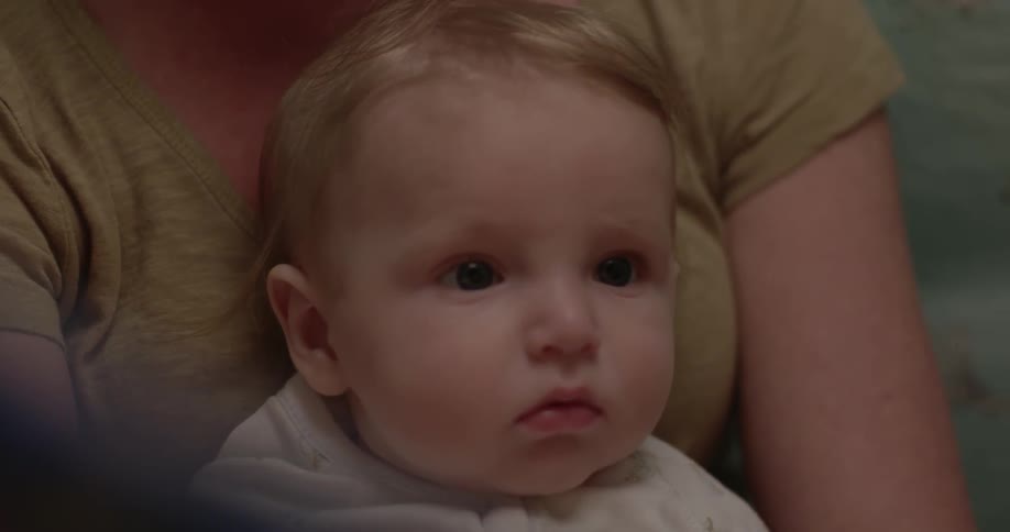 A baby sits on a woman's lap and looks at a book as she turns the pages