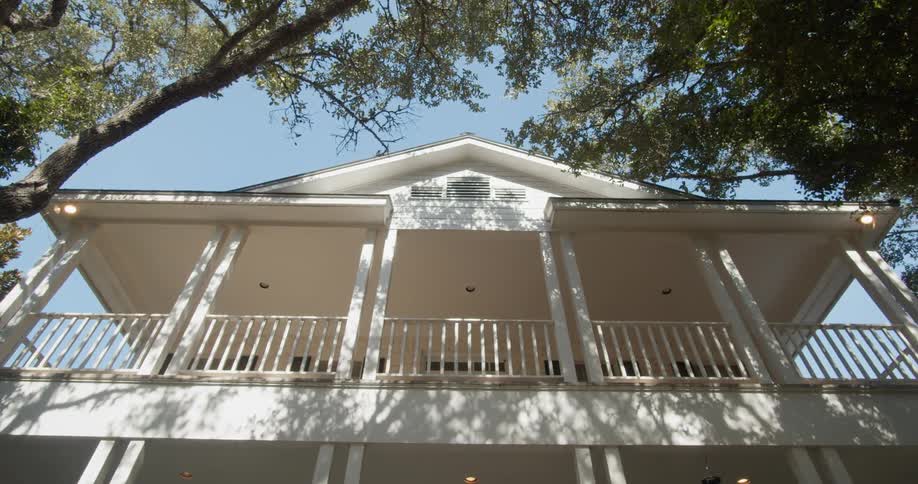 A large white house with a wide balcony is shaded by several tall trees