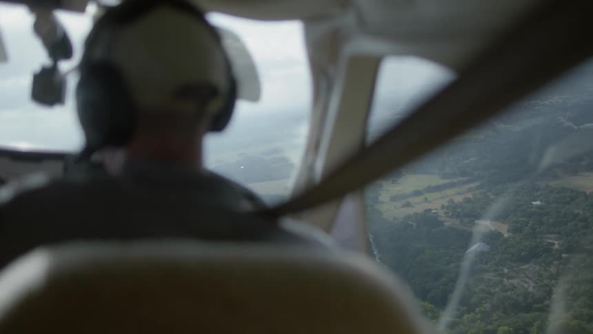 A man wearing headphones is in the copilot seat of a small airplane looking out the window at the land below