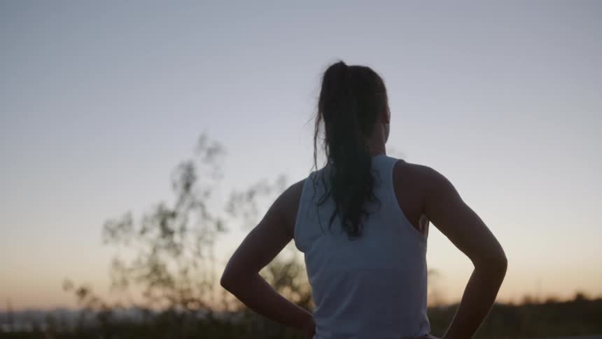 A woman in a white tank top walks outside at dusk