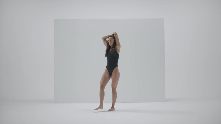 A woman wearing a bathing suit stands in an empty room stretching