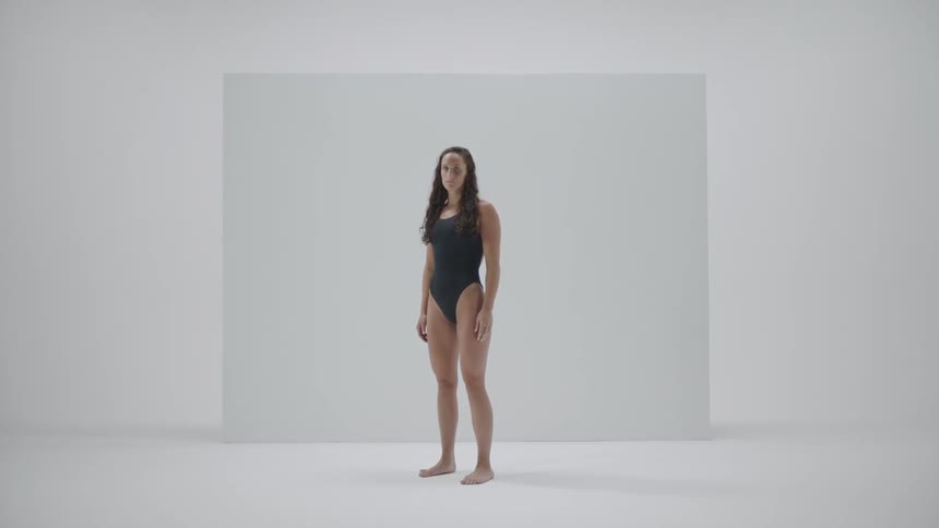 A dark haired young woman in a dark one piece bathing suit stands in front of a white wall