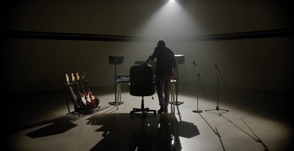 A spotlight illuminates a desk with a computer on it next to a rack of guitars