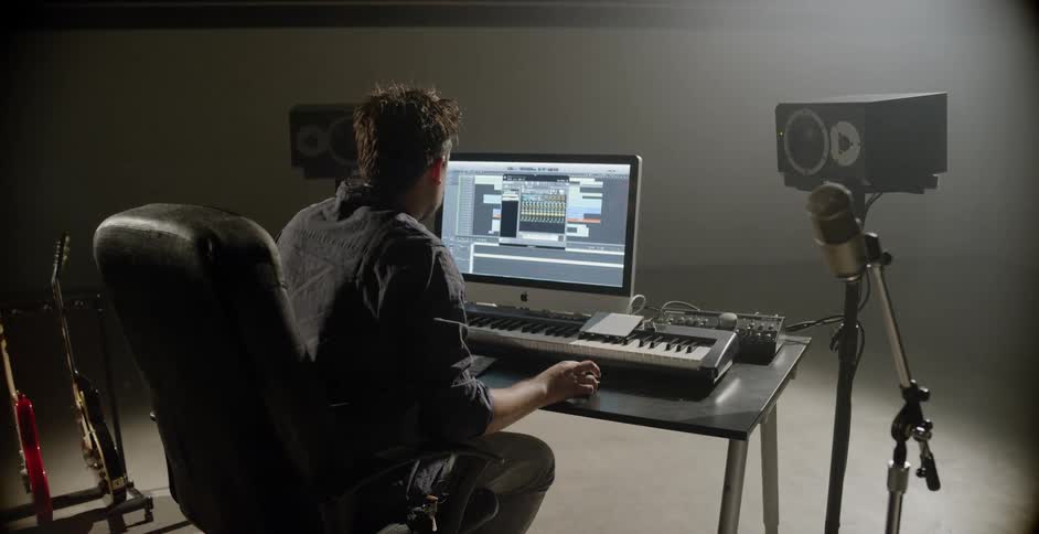 A man sits at a workstation with a monitor and a musical keyboard speakers on stands and microphone