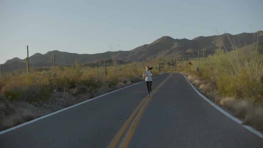 A female with long dark hair is running down the middle of a two lane road with cacti on either side of her and mountains in the distance
