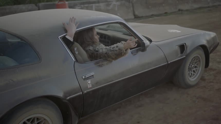 A woman in a fur jacket holds her arm out the window of a dusty car traveling on a dirt road