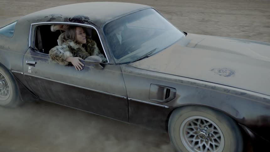 A woman sits in the front passenger seat of a classic sports car that is covered in dirt