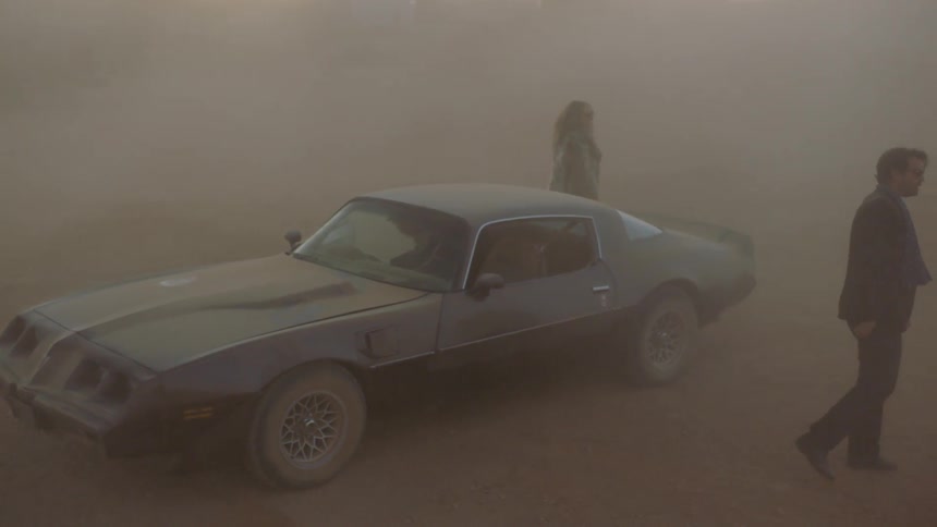 Three people get out of a car and walk away ina dusty field