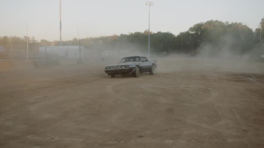 A vintage sports car is doing donuts in dusty dirt of a sports arena