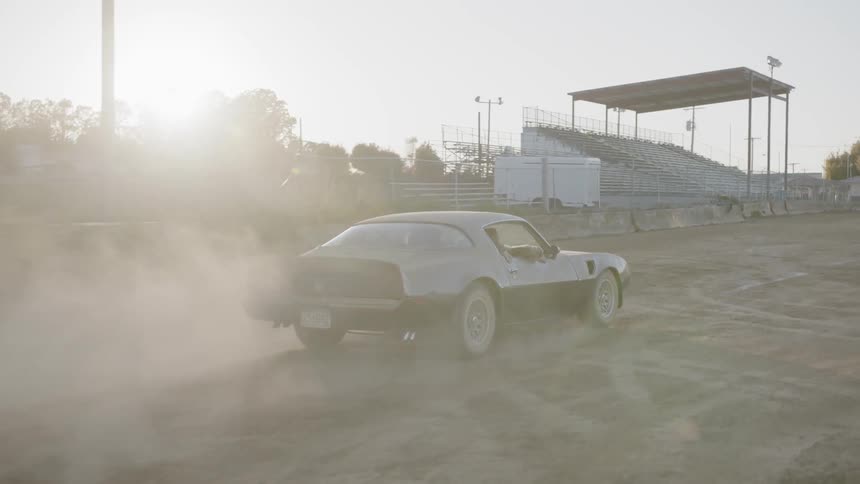 A classic sports car drives onto a dirt field that is surrounded by bleachers and a grandstand