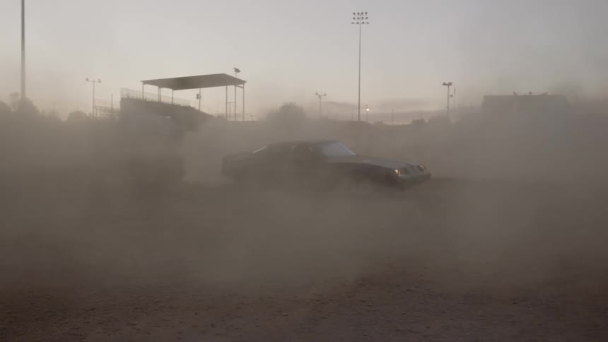 A classic muscle car is driving in circles in a large open dirt field while kicking up a large cloud of dust
