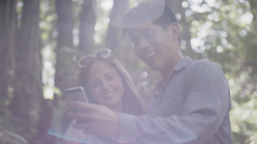 A man and a woman smile as they stand together in a forest and the man holds out his phone to take a selfie
