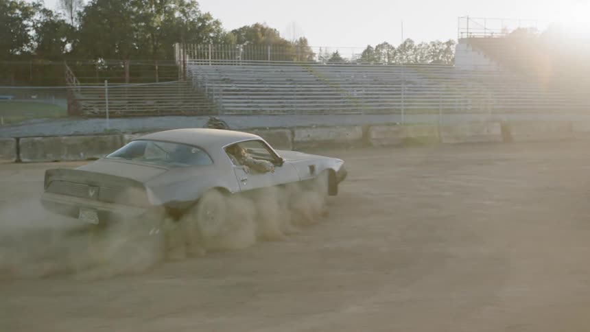 A black car kicking up a cloud of dust as it sharply turns within a dirt field surrounded by bleachers