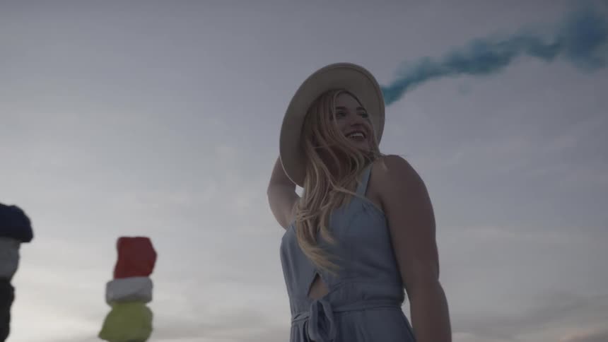 A young woman playfully releases blue smoke from a small canister above her head while standing near some stacked and painted rocks