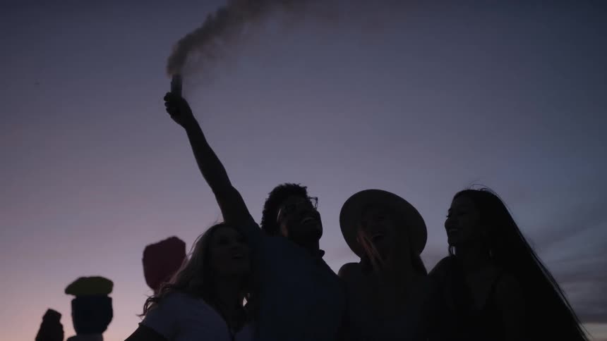 The silhouette of a group of young people can be seen against the evening sky as one releases smoke from a small cylinder above their heads