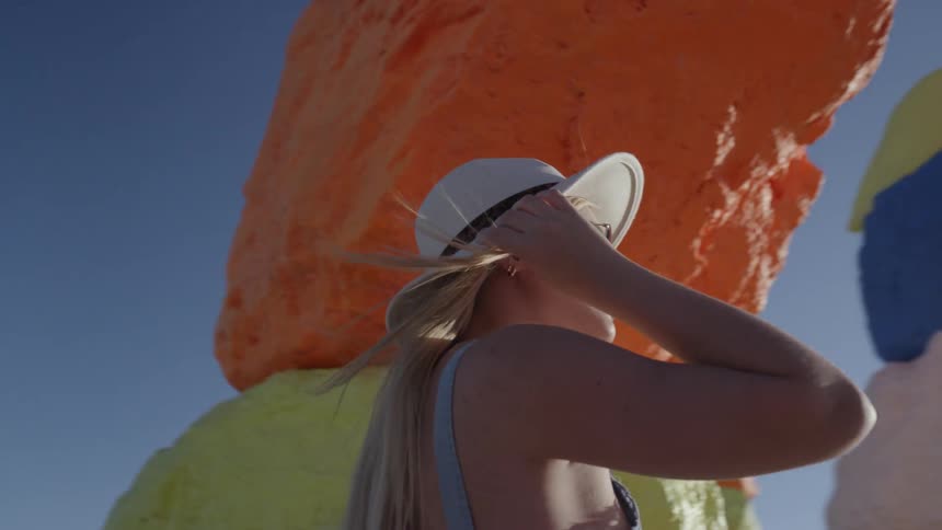 A young woman wearing a hat and sunglasses stands next to a large rock formation on a very sunny day