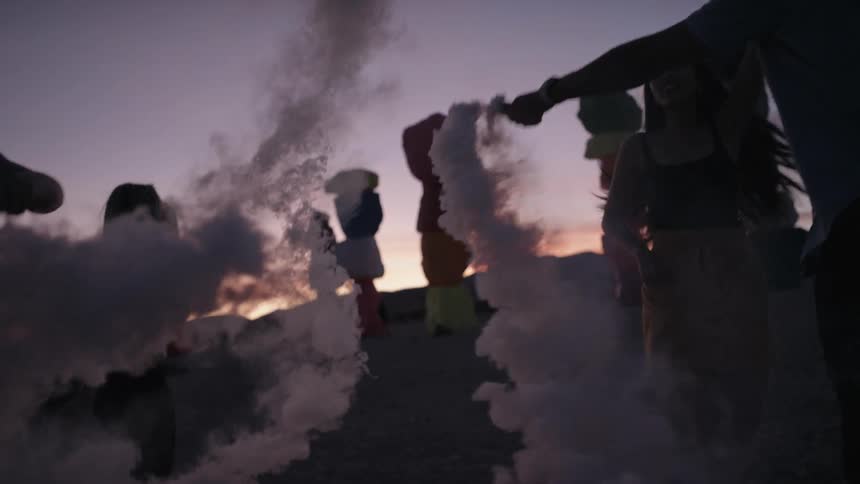 A group of people dance outside and wave a stick in the air as powder releases from it