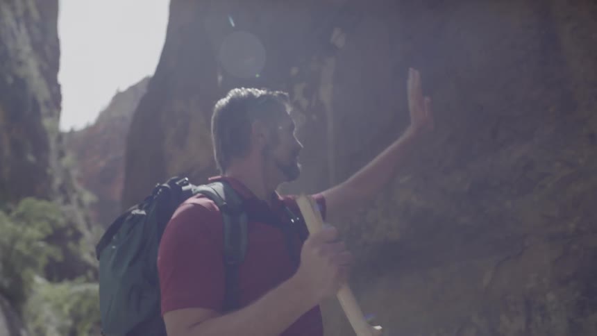 Man holding walking staff wades through river and touches cliff side