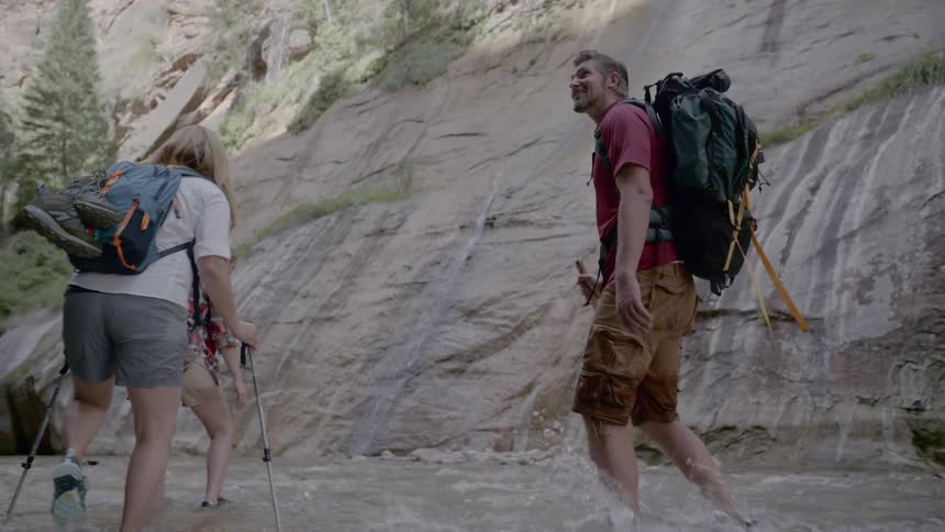Four people wearing backpacks are walking through a shallow stream with steep rocky cliffs beside them and two are using walking sticks