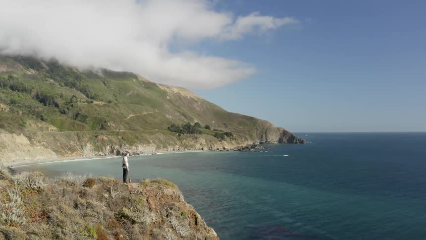 A white man stands on a cliff overlooking the ocean