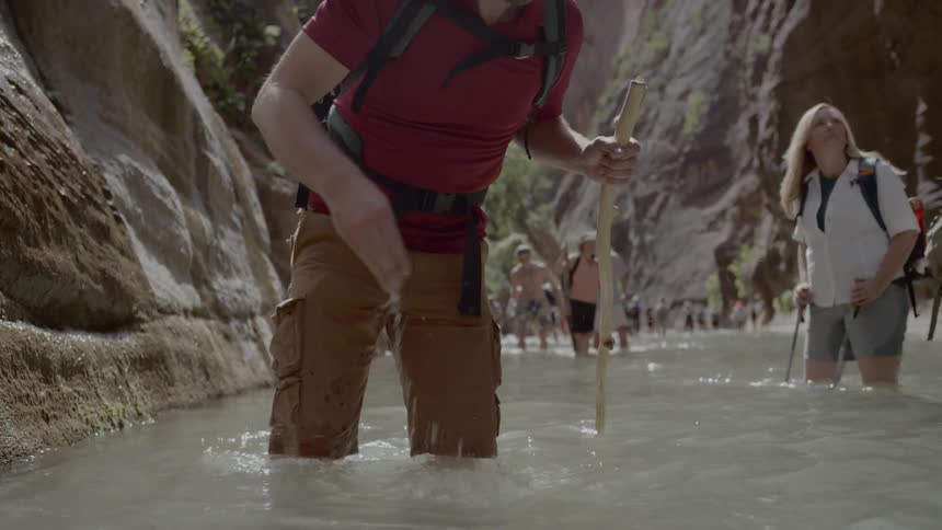 A group of people walk along the stream and a man drinks water from his palm between two mountains