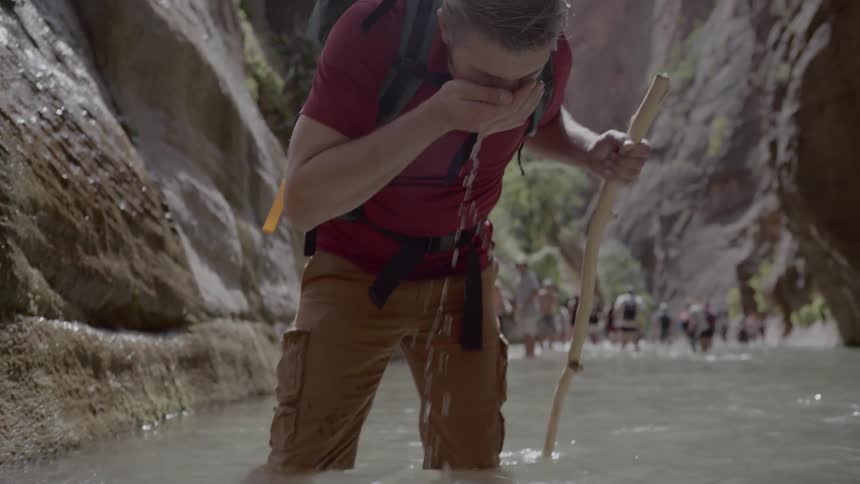 A group of people walk along the stream and a man drinks water from the stream with his palm between the two mountains