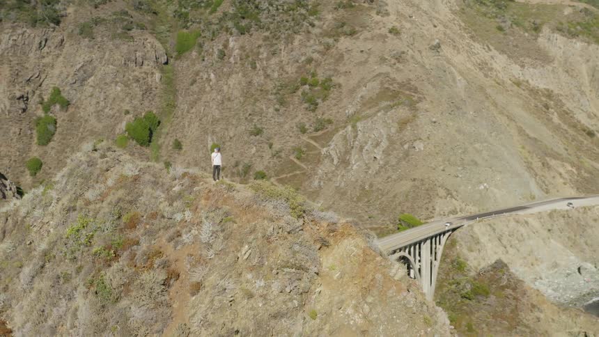 A person standing on a cliff near the coast with a bridge in the background