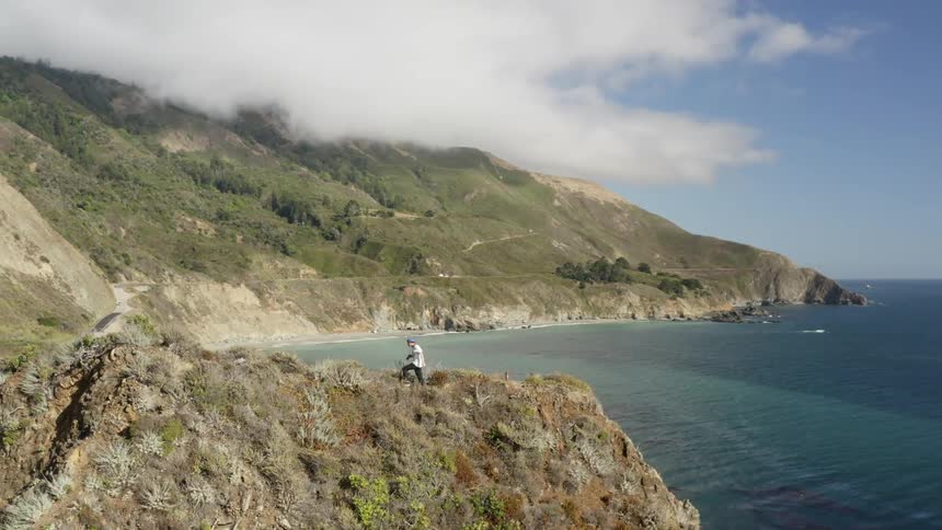 A man walks along the ridge of a hill along the ocean to get a vantage point to take pictures from
