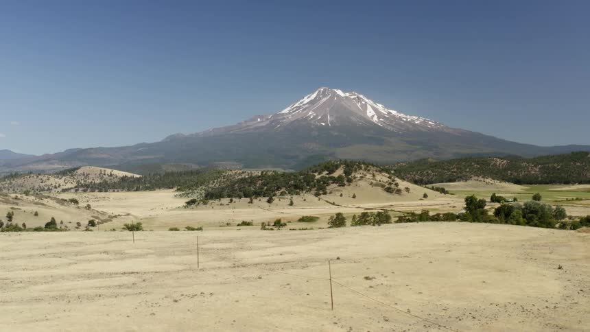 A snow capped peak against a blue sky with trees and sand and brush in the foreground