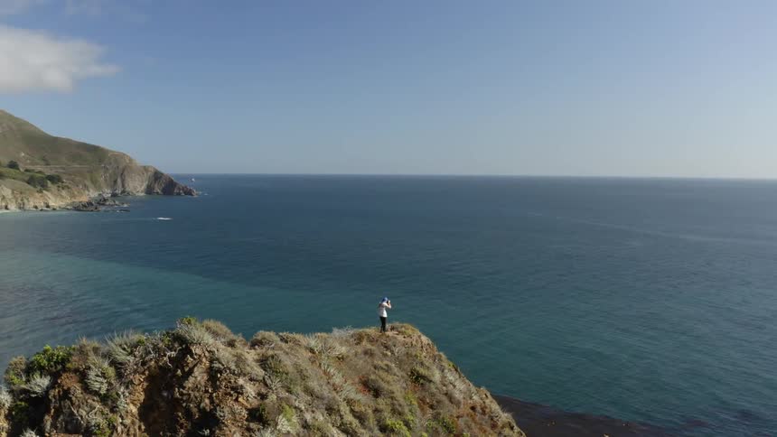 A person is standing on top of a mountain taking pictures of the ocean