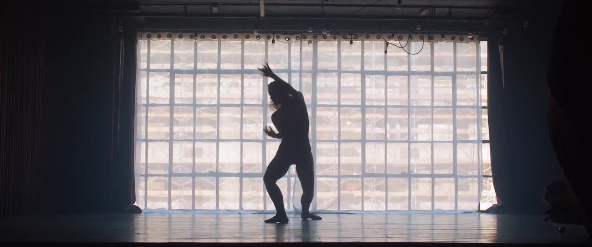 A woman dances on a stage with curtain covered windows behind her