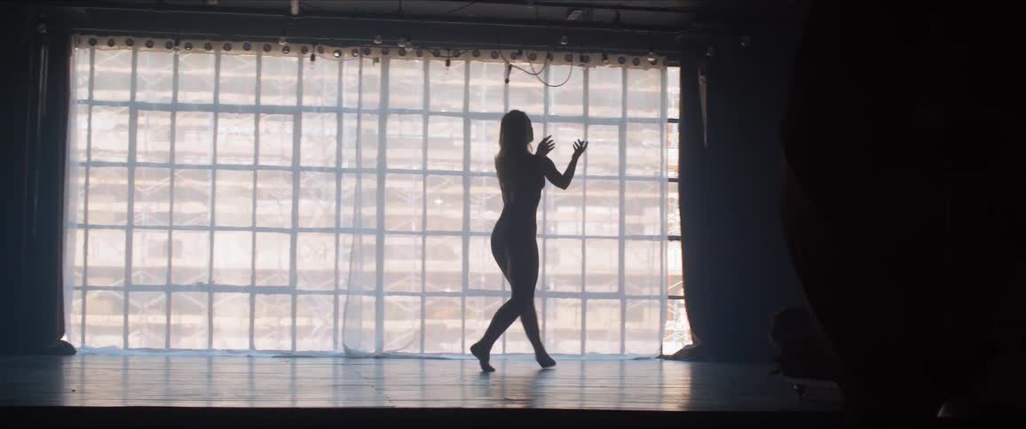 A female dancer is practicing on a stage in front of a full length mirror and wall of windows