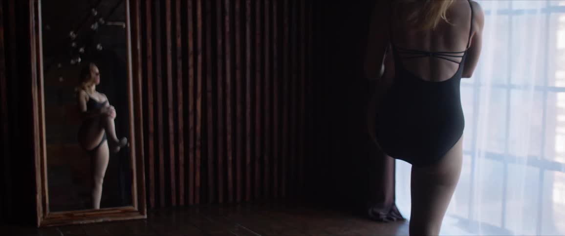A female dancer stretches her leg by pulling her heel up behind her and then knee to chest and her reflection can be seen in a full length mirror