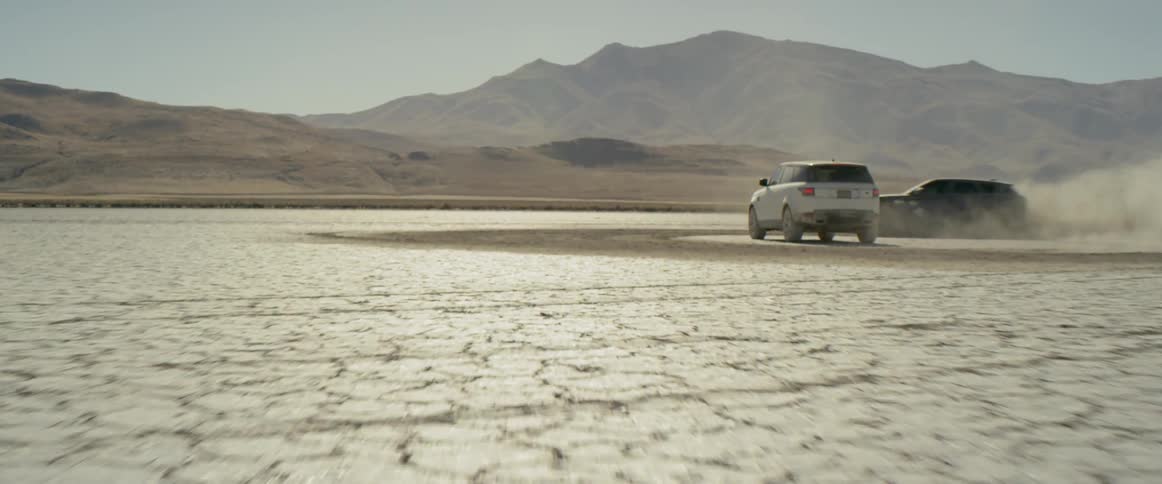 A car drives around a parked car on a dry and cracked dirt surface