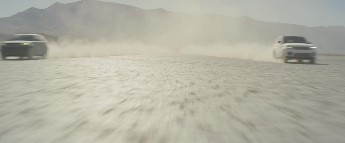 Two vehicles racing in the desert raising dust with mountains and the sky in the background