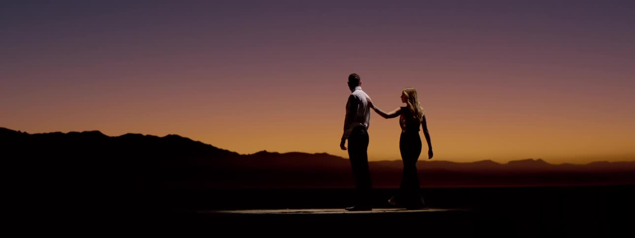 A man and woman standing in front of a sunset approach each other as they prepare to dance