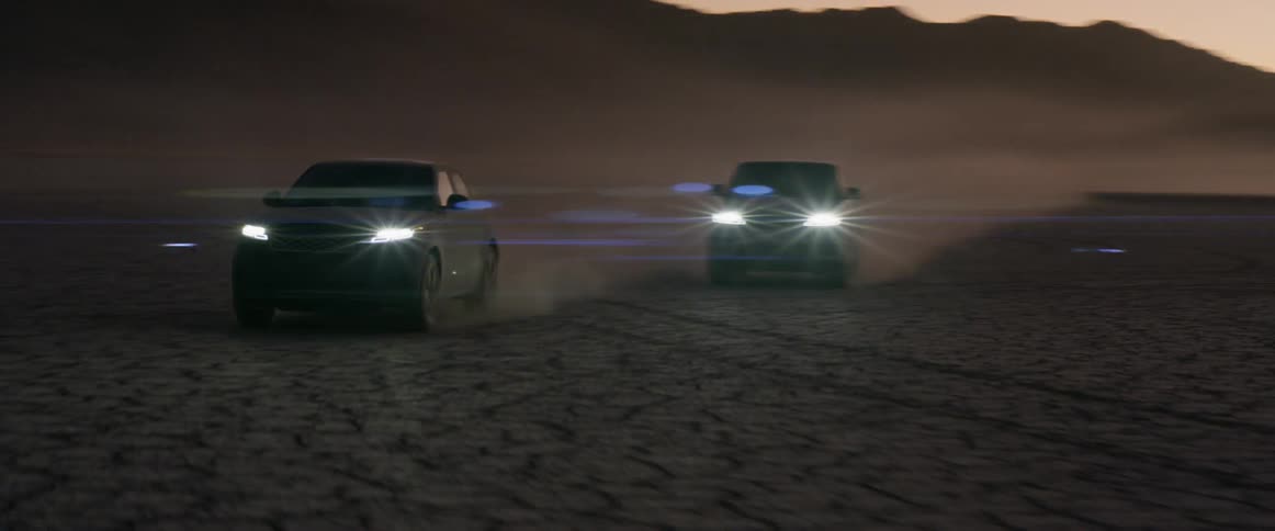 Two sport utility vehicles drive along dusty wide open landscape with their headlights on as they sun has just set