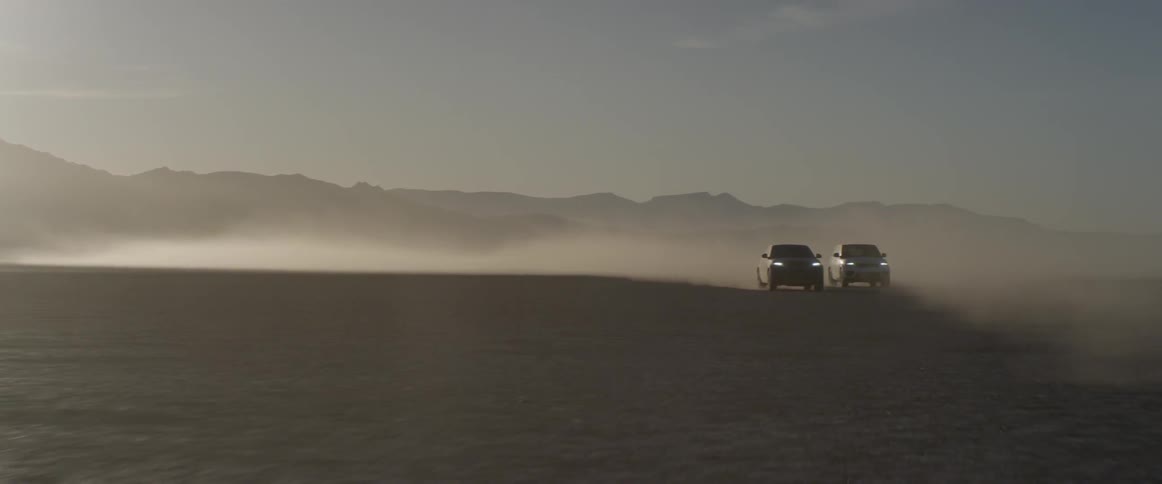 Two sport utility vehicles speed along next to each other through a flat dirt landscape
