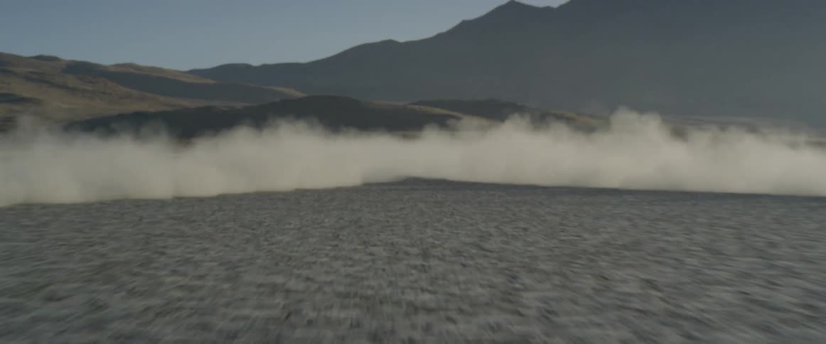 Two cars approach quickly across the open desert creating a dust cloud