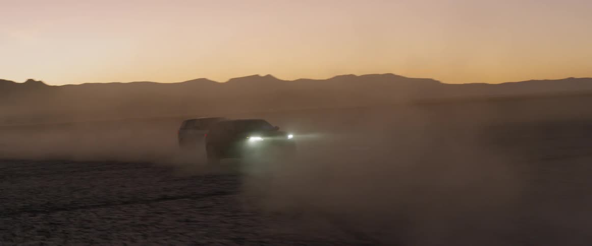 Sports utility vehicles race through dust clouds on desert near mountains after dusk