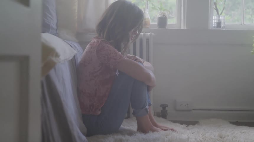 A woman hugging her knees while sitting on the floor with her back against a bed
