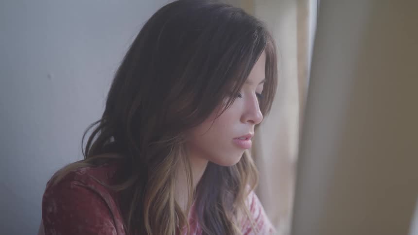 Sad young woman looking down sitting near a window indoors