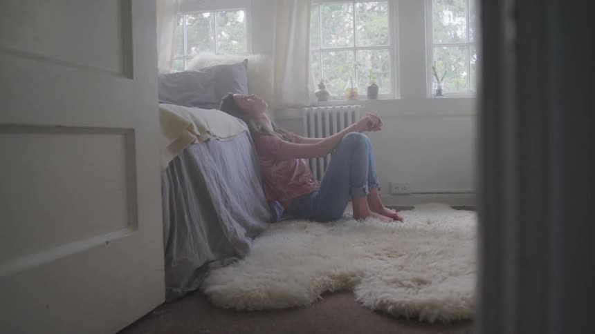 A girl sits on the floor of her room with her head leaning back against her bed