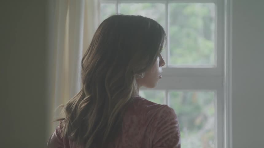 A woman with long brown hair stands next to a window
