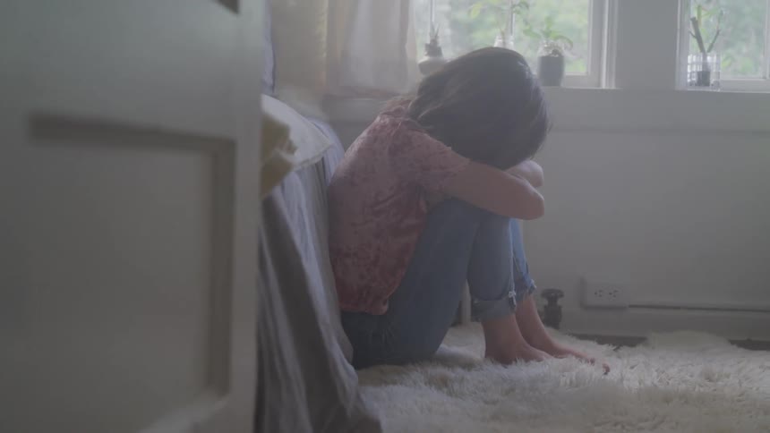 A young girl sits on her bedroom floor with her head bowed and knees pulled to her chest