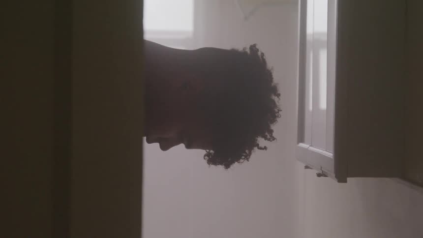 A young black male sits in a small room while hanging his head downward
