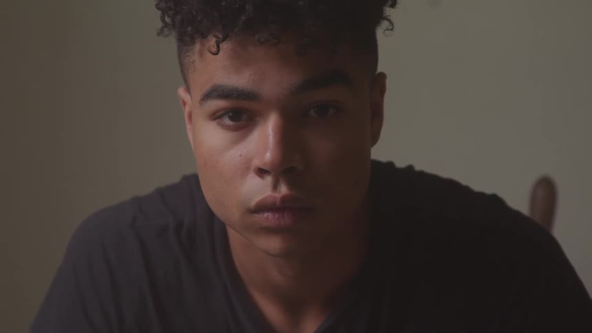 A young black man sitting down in a room stares straight ahead