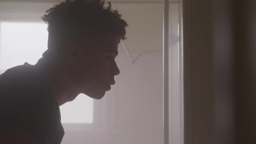 A young man stands in a bathroom and looks at himself in the mirror