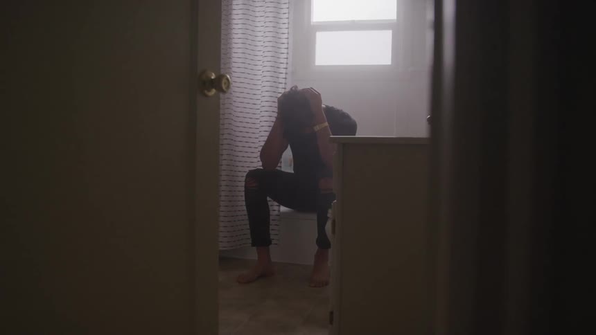 A young man sits on the edge of a bathtub with his head in his hands