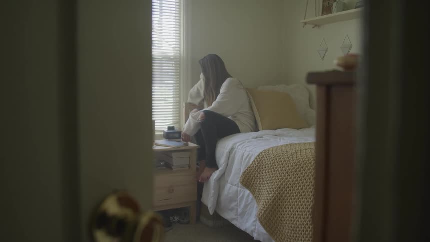 A young girl sits on her bed and looks out the window